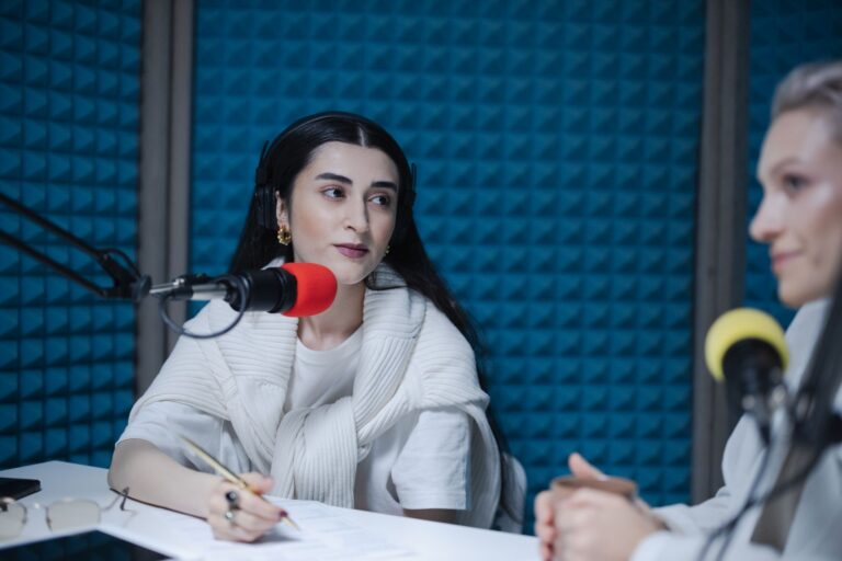 Două persoane care discută la o masă de bucătărie, cu un reportofon și un carnet de notițe între ele